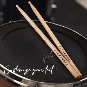 May include: A pair of wooden drumsticks, each engraved with the name "ALEXANDER", lying on a black drum. The image includes the text "Customize your text" in a cursive font.