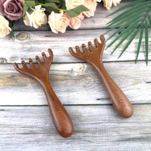 May include: Two wooden head massagers with a fork-like design. The massagers are a rich brown colour and have a smooth finish. The handles are engraved with the word "Vera". The background is a light wood surface with flowers.