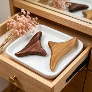 May include: Two wooden gua sha tools, one dark brown and one light brown, rest on a white ceramic tray. The tools are triangular with curved edges. The tray sits inside a wooden drawer, with a small bouquet of pink flowers in the background.