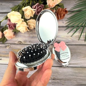 May include: A silver compact mirror and brush set. The brush has black bristles with white tips. A pink bow decorates the mirror's cover, with the text "Leblond Est. 7.11.2020" engraved below. The set is open, revealing the mirror.