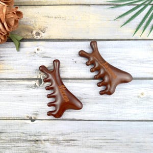 May include: Two dark brown wooden massage tools with a comb-like design. Each tool has a name engraved on it. One says "Steven" and the other says "James". The tools are on a light wood surface.