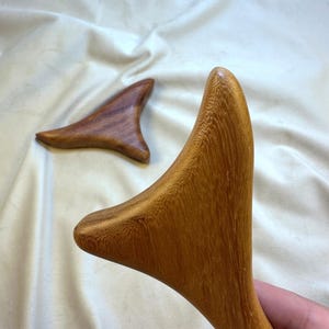 May include: Two wooden gua sha tools. One is in focus, showing a light brown color with a smooth, curved design. The other is in the background, with a darker brown color. The tools are on a white, textured surface.