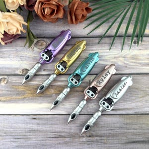 May include: Five personalized pens in various colors: purple, gold, teal, rose gold, and silver. Each pen has a silver tip and a name engraved on the barrel: Erik, Thomas, Calvin, Ryley, and Xavien. The pens are arranged on a wooden surface.