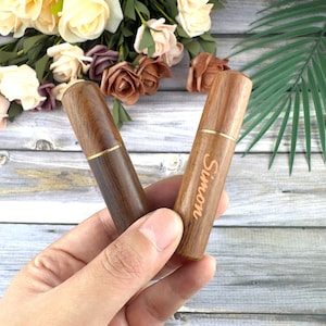 May include: Two wooden needle cases, one with the name "Simon" engraved. The cases are cylindrical with a gold-colored band near the top. The wood grain varies in color from light to dark brown. The background includes flowers.