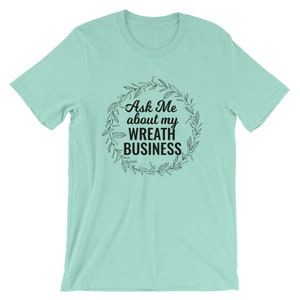 Peut inclure: Un t-shirt bleu clair avec un motif de couronne noire et le texte "Ask Me about my Wreath Business".
