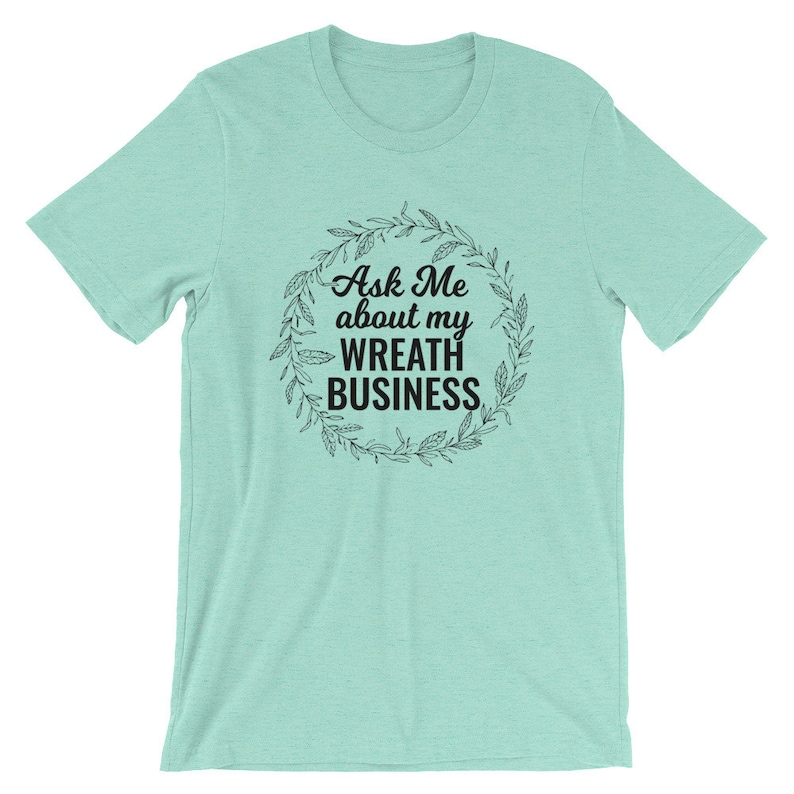 Peut inclure: Un t-shirt bleu clair avec un motif de couronne noire et le texte "Ask Me about my Wreath Business".