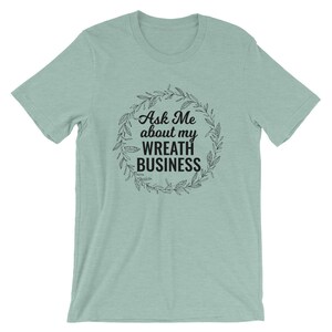Peut inclure: Un t-shirt bleu clair avec un graphique noir d'une couronne avec le texte "Ask Me about my Wreath Business".