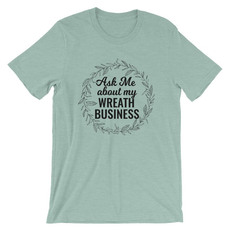 Peut inclure: Un t-shirt bleu clair avec un graphique noir d'une couronne avec le texte "Ask Me about my Wreath Business".