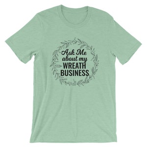 Peut inclure: Un t-shirt vert clair avec un motif noir qui dit "Ask Me about my Wreath Business" &agrave; l'int&eacute;rieur d'une couronne de feuilles.