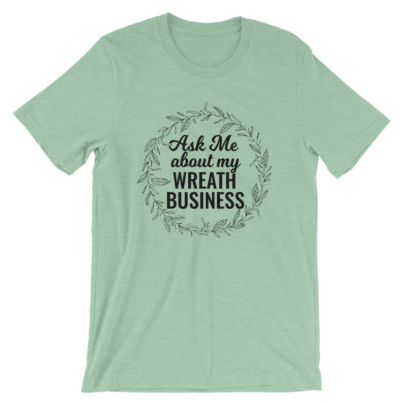 Peut inclure: Un t-shirt vert clair avec un motif noir qui dit "Ask Me about my Wreath Business" &agrave; l'int&eacute;rieur d'une couronne de feuilles.