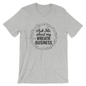 Peut inclure: Un t-shirt gris chin&eacute; avec un motif noir qui dit "Ask Me about my Wreath Business" entour&eacute; d'une couronne de feuilles.