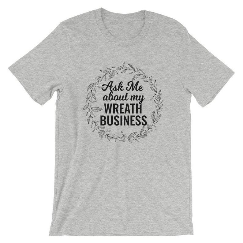 Peut inclure: Un t-shirt gris chin&eacute; avec un motif noir qui dit "Ask Me about my Wreath Business" entour&eacute; d'une couronne de feuilles.
