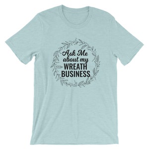 Peut inclure: Un t-shirt bleu clair avec le texte "Ask Me about my Wreath Business" &agrave; l'int&eacute;rieur d'une couronne.