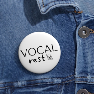 Könnte beinhalten: Ein weißer Ansteckbutton mit dem schwarzen Schriftzug "VOCAL rest" und einem durchgestrichenen Mikrofon-Symbol. Der Button ist auf einer blauen Jeansjacke befestigt.