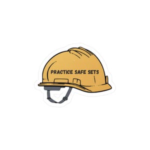 May include: A yellow hard hat sticker with the text "PRACTICE SAFE SETS" in black. The sticker has a black outline and a gray chin strap. The hard hat is a common safety item used in construction and other industrial settings.