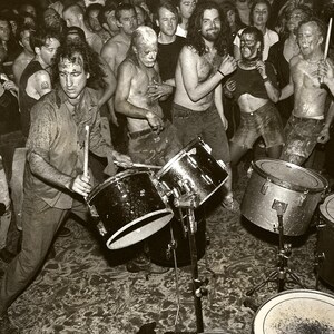 May include: A black and white photo of a group of people playing drums in a crowded room. The drums are set up on a stage and the people are standing around them, playing and singing. The photo is taken from a low angle, looking up at the stage.