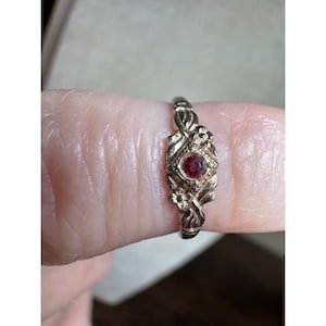 May include: A gold-toned metal ring featuring a central round red gemstone. The band is decorated with embossed floral motifs and textured leaf-like patterns surrounding the stone, creating a detailed and ornate appearance on the ring surface.