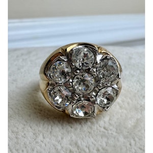May include: A gold-toned ring featuring a cluster of seven clear, round-cut gemstones. The gemstones are set in a circular pattern, creating a floral or starburst design. The ring has a thick band and a polished finish.