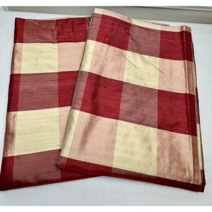 May include: Two red and cream-colored plaid curtains. The fabric has a subtle sheen, and the pattern is a classic check design. The curtains are folded, showing the full pattern. The color scheme is a mix of deep red and a creamy off-white.