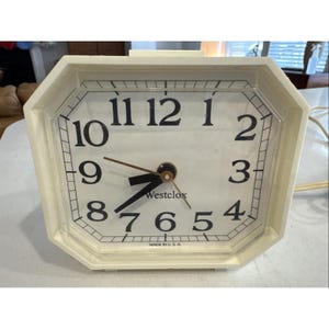 May include: Cream-colored Westclox electric clock with a rectangular face and octagonal frame. Black numerals mark the hours on the white clock face. The clock hands are black and brown. The words "Westclox" and "MADE IN U.S.A." are printed on the face.