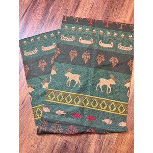 May include: A woven green throw blanket featuring a repeating pattern of tan moose, brown maple leaves, yellow pine trees, red fish, and small canoes. The design includes horizontal bands of various nature-themed motifs in earthy tones.