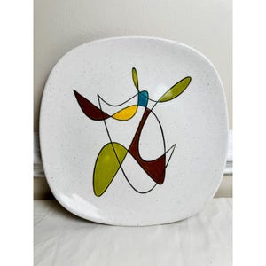 May include: White square plate with a speckled surface. The plate features an abstract design with black outlines and colorful shapes in yellow, green, teal, and maroon. The plate has rounded corners.