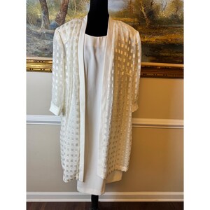 May include: A two-piece ensemble consisting of a sleeveless, solid off-white dress and a matching long-sleeved, open-front duster jacket. The jacket features a sheer, textured fabric with a repeating grid pattern of small, opaque white squares.
