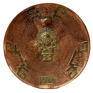 May include: A round copper-colored metal plate with a central gold-colored figure and geometric designs. The word "PERU" is inscribed at the bottom. The plate has a textured surface and a decorative design.