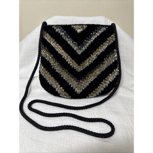 May include: A black velvet crossbody bag with a chevron pattern of silver and gold beads. The bag has a black rope strap and a curved shape. The bag is approximately 8 inches wide and 7 inches tall.