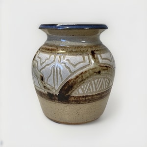 May include: A handcrafted ceramic vase with a wide base and a slightly flared rim. The vase features a light tan base, a band of white and brown patterned design, and a dark blue rim. The overall aesthetic is rustic and artistic.