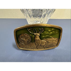 May include: A rectangular, vintage belt buckle with a gold-toned frame and a green enamel center. The buckle features a detailed depiction of a stag with large antlers, set against a landscape. The buckle measures approximately 3 inches in length.