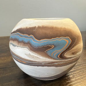 May include: A round, ceramic vase with a marbled design. The vase features a blend of colors, including white, brown, blue, and orange, creating a unique, swirling pattern. The vase is sitting on a wooden surface.