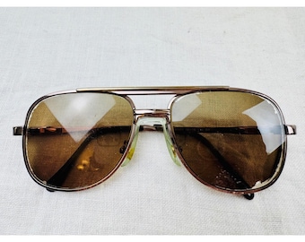 Puede incluir: Gafas de sol tipo aviador con montura de bronce metálico, diseño de doble puente y lentes tintadas en marrón degradado. Las gafas tienen una forma clásica de lágrima con patillas finas y almohadillas nasales transparentes.