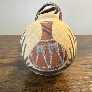 May include: A round, beige ornament with a brown leather strap. The ornament features a painted design of a brown vase with silver and black accents. Black vertical lines and leaf-like shapes surround the vase design.