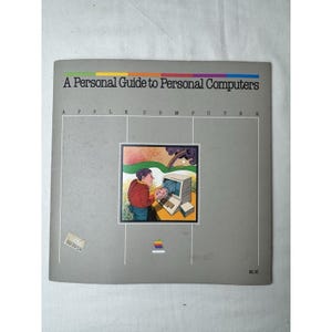 May include: A vintage book cover titled "A Personal Guide to Personal Computers." The cover is gray with a colorful stripe at the top. It features an illustration of a person using an early Apple computer. The Apple logo is visible.