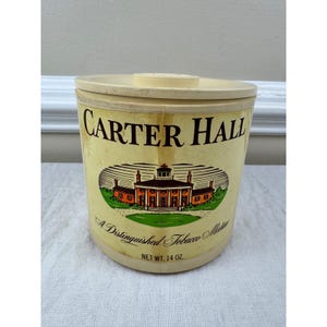 May include: A cylindrical Carter Hall tobacco container with a cream-colored lid and body. The label features the text CARTER HALL, an illustration of a large house, and the text A Distinguished Tobacco Mixture, NET WT. 14 OZ. in black and orange.