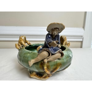 May include: A ceramic planter with a seated figure wearing a hat and blue robe. Two frog figurines sit on the planter's rim. The planter is green with brown accents.