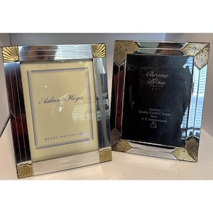 May include: Two rectangular photo frames with silver and gold accents. One frame has the text "Ashton & Hayes" and "MIXED METALS 5X7". The other frame has the text "Chrome & Brass" and "Another Quality Crafted Frame from A K International".