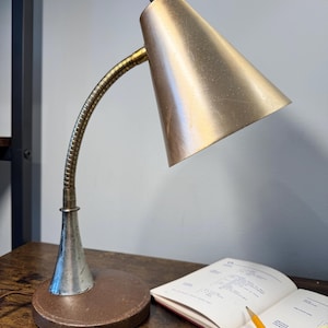 May include: A vintage desk lamp with a conical, gold-colored shade and a flexible, brass-toned neck. The lamp sits on a brown, circular base. A notebook and pencil are on the wooden desk surface.