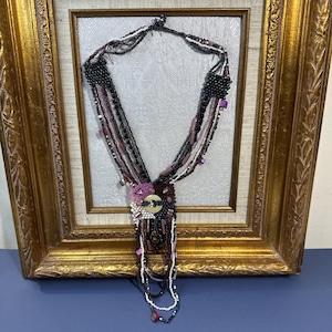 May include: A multi-strand beaded necklace with a variety of colors, including black, white, and shades of purple. The necklace features a central floral design with a large button and dangling beaded strands. The necklace is displayed in a gold framed picture.