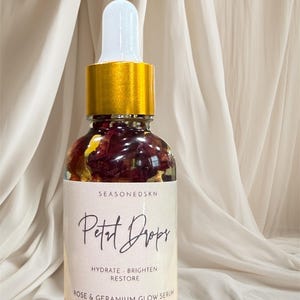 Könnte beinhalten: Eine klare Glasflasche mit SEASONEDSKN Petal Drops Rose & Geranium Glow Serum. Die Flasche hat einen goldenen Verschluss und eine weiße Pipette. Das Etikett lautet "Hydrate, Brighten, Restore" und enthält 30 ml Produkt.