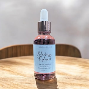 Könnte beinhalten: Eine durchsichtige Glasflasche mit Tropferaufsatz, gefüllt mit einer rosa Flüssigkeit. Die Flasche hat ein weißes Etikett mit dem Text "Blueberry Radiance Facial Serum 100% Natural Botanical Extracts 1oz | 30ml".
