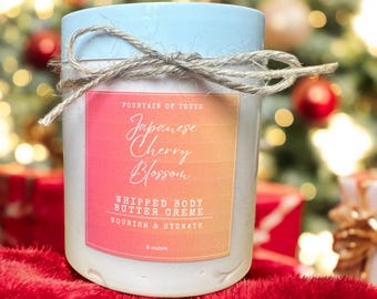 May include: A white jar with a light blue lid and a brown twine bow. The jar contains whipped body butter creme with a label that reads "Fountain of Youth Japanese Cherry Blossom Whipped Body Butter Creme Nourish & Hydrate 8 ounces".