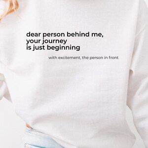 Może przedstawiać: Biała bluza z kapturem z czarnym napisem: "dear person behind me, your journey is just beginning", z napisem "with excitement, the person in front" poniżej. Bluza z kapturem jest noszona z jasnoniebieskimi dżinsami.