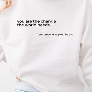 Peut inclure: Un sweat à capuche blanc avec le texte "you are the change the world needs" et "from someone inspired by you" imprimé en noir. Le sweat à capuche a une capuche et des manches longues. Le modèle porte un jean bleu clair.