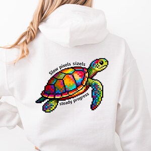 Könnte beinhalten: Weißer Kapuzenpullover mit einem farbenfrohen, pixeligen Schildkrötendesign. Die Schildkröte hat einen lebendigen Panzer mit Rot-, Gelb-, Blau- und Grüntönen. Der Text "Slow pixels sizzle, steady progress" befindet sich über und unter der Schildkröte.
