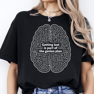 Puede incluir: Camiseta negra con un diseño de laberinto cerebral blanco y el texto "Getting lost is part of the genius plan". El diseño está centrado. La camiseta tiene mangas cortas y cuello redondo.