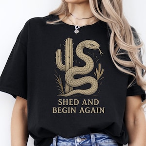 Può includere: T-shirt nero con un design a tema deserto. Il design include un serpente arrotolato, un cactus e il testo "SHED AND BEGIN AGAIN" in oro. È un capo d'abbigliamento casual.
