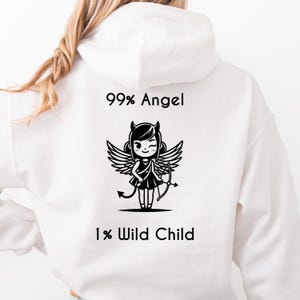 Puede incluir: Sudadera con capucha blanca con el texto "99% Angel 1% Wild Child" encima y debajo de un gráfico en blanco y negro de un ángel guiñando un ojo con cuernos de diablo, alas y arco y flecha.