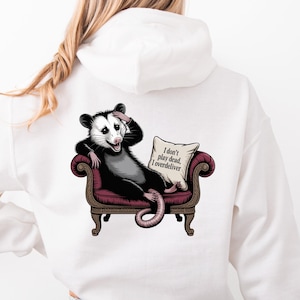 May include: White hoodie featuring a cartoon opossum lounging on a maroon couch, with the text "I don't play dead, I overdeliver" on a pillow. The opossum is black, white, and gray.